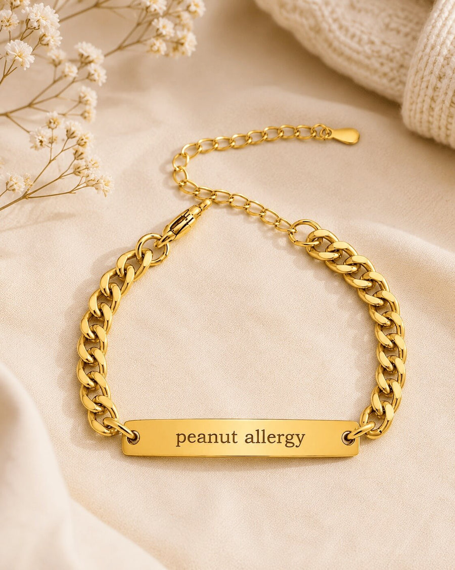 Gold chain bracelet with 'peanut allergy' engraving on a beige fabric background