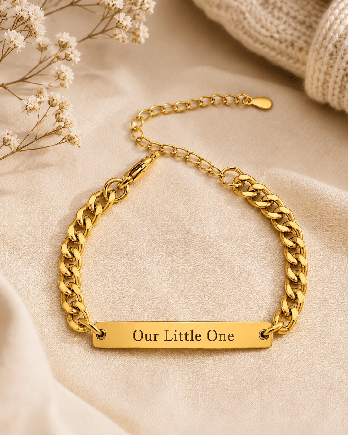 Gold chain bracelet with 'Our Little One' engraving on a beige fabric background